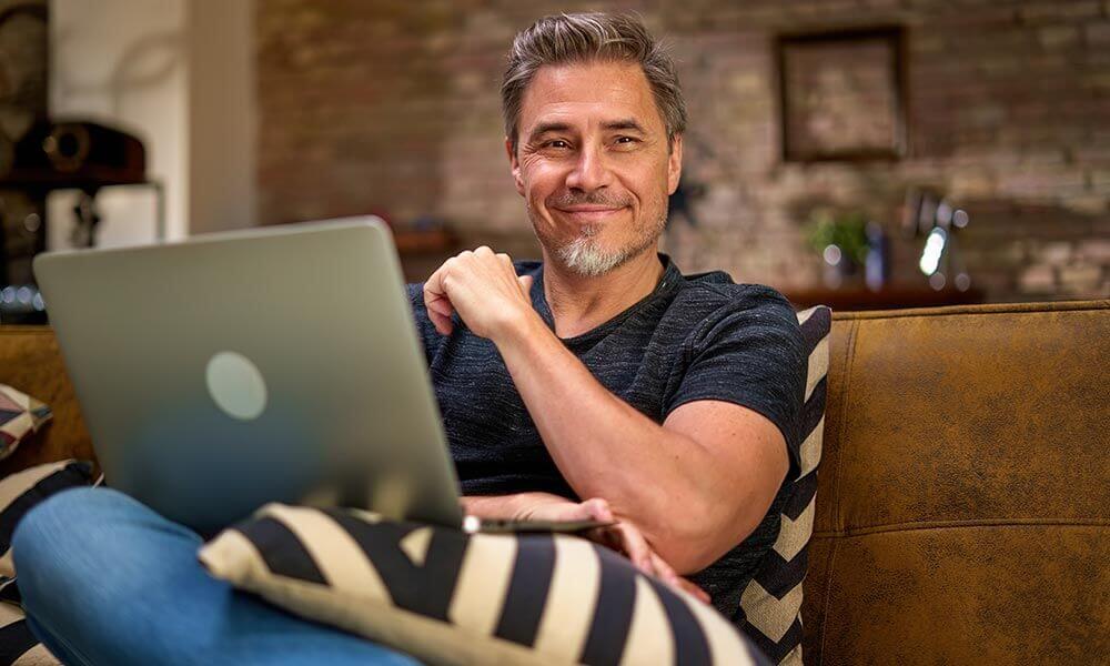 Man Smiling While Using His Laptop Computer on a Couch